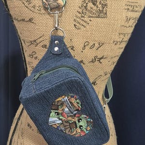 May include: Denim sling bag with a green strap and zipper. The bag features a colorful patch with cartoon characters. The strap is attached to a decorative patch with a colorful design.
