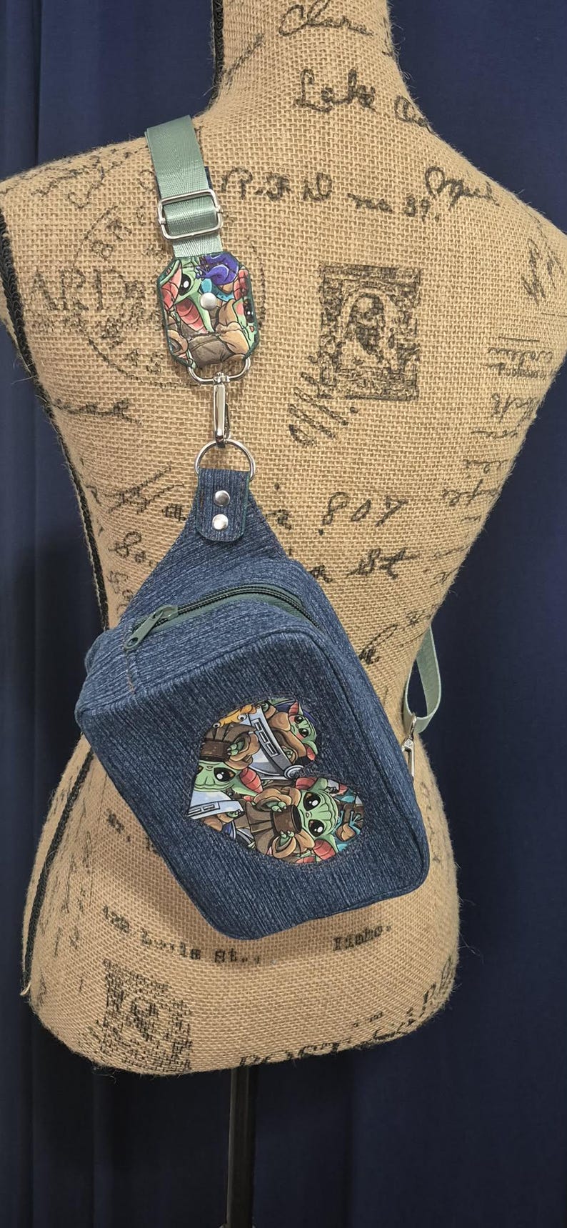 May include: Denim sling bag with a green strap and zipper. The bag features a colorful patch with cartoon characters. The strap is attached to a decorative patch with a colorful design.