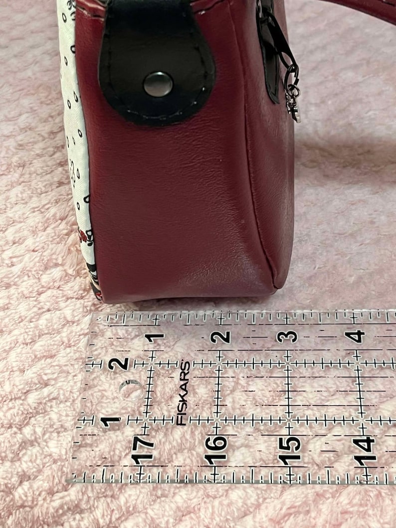 Puede incluir: Primer plano de un bolso de cuero rojo con un patr&oacute;n de lunares blancos y negros. El bolso se mide con una regla, mostrando que tiene aproximadamente 7,6 cm de ancho.