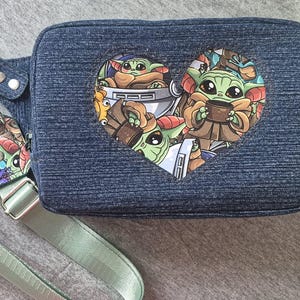 May include: A denim blue fanny pack with a heart-shaped graphic featuring the character Grogu from The Mandalorian. The pack has a light green adjustable strap and a matching keychain. The bag is approximately 10 inches wide.