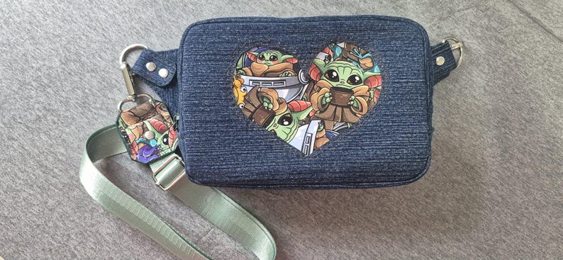 May include: A denim blue fanny pack with a heart-shaped graphic featuring the character Grogu from The Mandalorian. The pack has a light green adjustable strap and a matching keychain. The bag is approximately 10 inches wide.