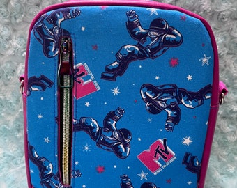 May include: A pink and blue crossbody bag with a zippered pocket and a repeating pattern of astronauts and the MTV logo. The bag has a pink strap and a blue and white fabric lining.