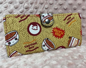 May include: A yellow and red fabric wallet with a ramen noodle pattern. The wallet has a snap closure and features the text "Caution Hot", "Rich Flavor", and "Maruchan Instant Lunch".