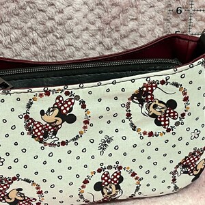 Puede incluir: Un bolso de hombro burdeos y negro con un forro de tela blanco con un patr&oacute;n repetido de Minnie Mouse en una corona de flores.