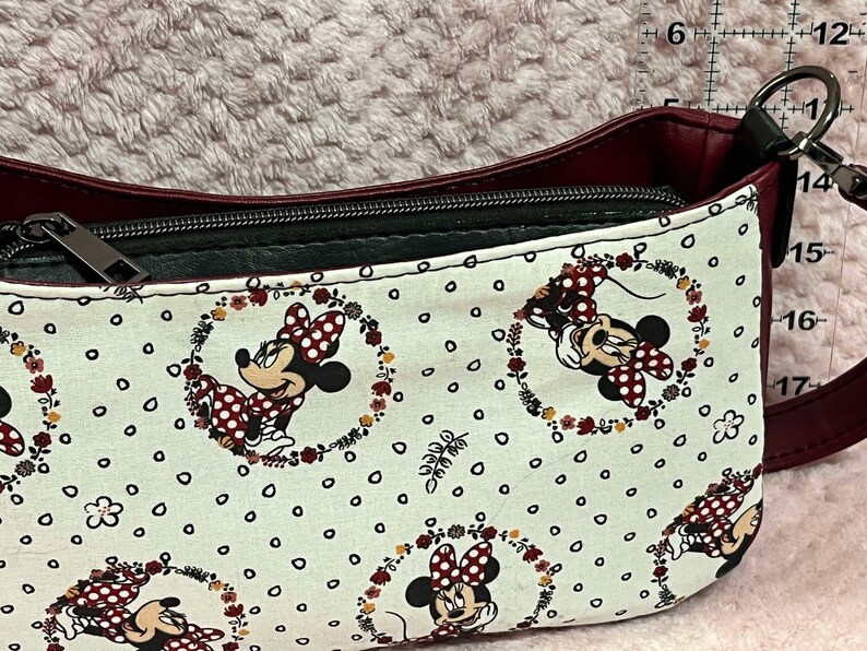 Puede incluir: Un bolso de hombro burdeos y negro con un forro de tela blanco con un patr&oacute;n repetido de Minnie Mouse en una corona de flores.
