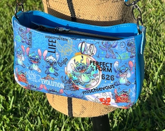 May include: A blue crossbody bag with a zipper closure and a colorful print featuring Stitch from Lilo & Stitch. The print includes the words "Life", "Indestructible", "Origin", "Evolution", "Perfect Form", "626", and "Mischievous".