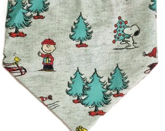 May include: A light gray dog bandana with a repeating pattern of cartoon Christmas trees and characters. The characters include Charlie Brown, Snoopy, and Woodstock. The trees are teal with red bases. The bandana is triangular in shape.