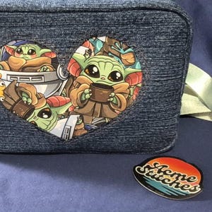 May include: A denim-textured lunch bag with a heart-shaped graphic featuring a cartoon character. The bag has a light green strap and a round pin with the text "Home Stitches".