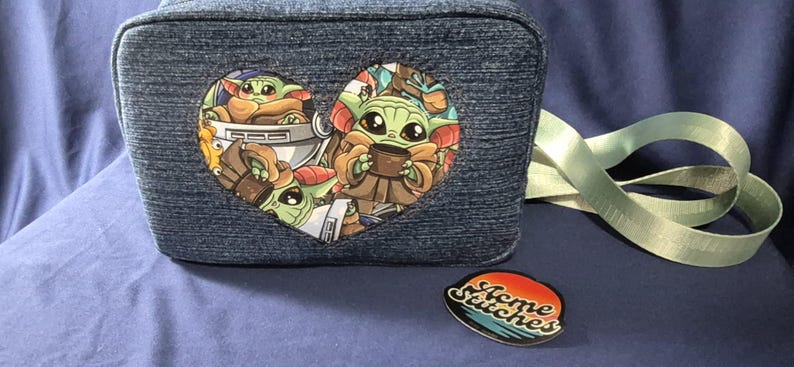 May include: A denim-textured lunch bag with a heart-shaped graphic featuring a cartoon character. The bag has a light green strap and a round pin with the text "Home Stitches".