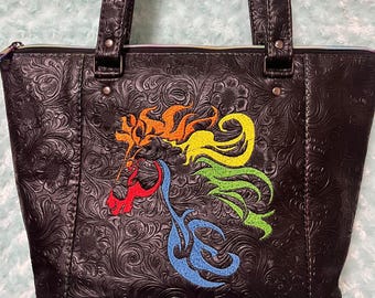 May include: A brown leather tote bag with a floral embossed design. The bag has a zipper closure and two top handles. The front of the bag features a colorful embroidered design of a stylized horse head.