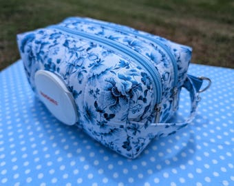 May include: A rectangular quilted fabric pouch featuring a blue and white floral pattern. The pouch has two zippered compartments, a side carrying strap with a metal ring, and a white plastic flip-top dispenser lid on the front side.