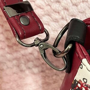 Puede incluir: Primer plano de una correa de bolso de cuero rojo con un cierre de metal plateado. La correa est&aacute; unida a un asa de cuero negro.