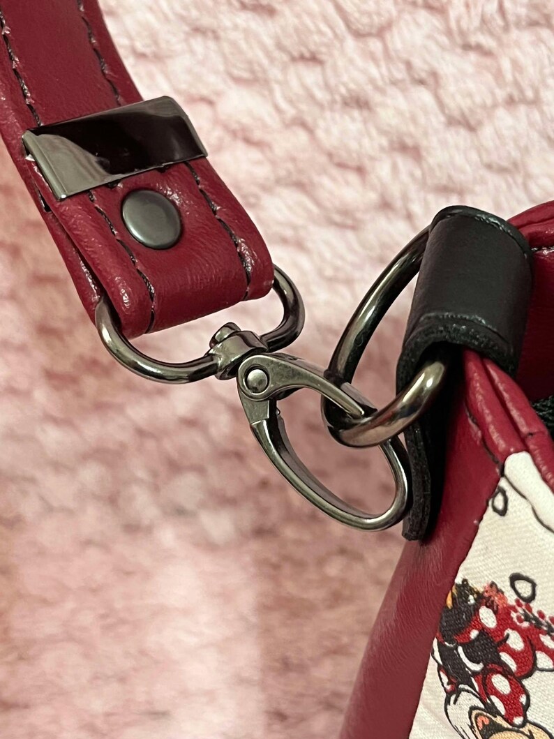 Puede incluir: Primer plano de una correa de bolso de cuero rojo con un cierre de metal plateado. La correa est&aacute; unida a un asa de cuero negro.