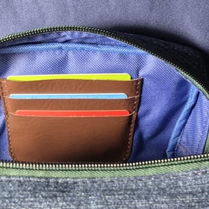 May include: A brown leather card holder with three colorful cards inside, nestled within a denim-textured pouch. The pouch has a blue interior and a green zipper, set against a blue background.