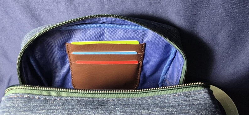 May include: A brown leather card holder with three colorful cards inside, nestled within a denim-textured pouch. The pouch has a blue interior and a green zipper, set against a blue background.