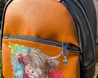 May include: A brown and black faux leather backpack with a colorful embroidered Highland Cow design. The backpack has a zipper closure and a small heart-shaped charm.