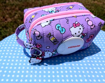 May include: A quilted purple rectangular pouch featuring a repeating Hello Kitty and candy print. The bag has a pink zipper, a side carrying strap with a Hello Kitty charm, and a white circular Huggies dispenser lid integrated into the front.