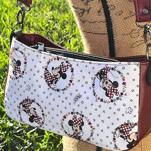 Puede incluir: Un bolso bandolera rojo y blanco con estampado de Minnie Mouse. El bolso tiene cierre de cremallera y una correa larga ajustable.