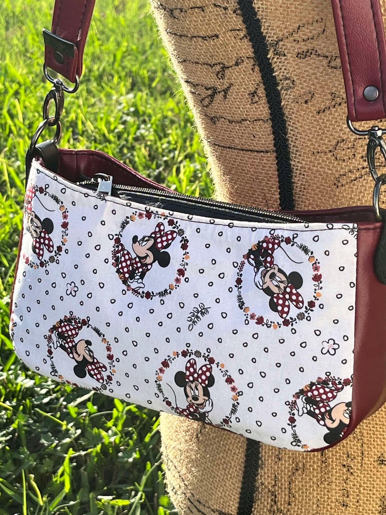 Puede incluir: Un bolso bandolera rojo y blanco con estampado de Minnie Mouse. El bolso tiene cierre de cremallera y una correa larga ajustable.