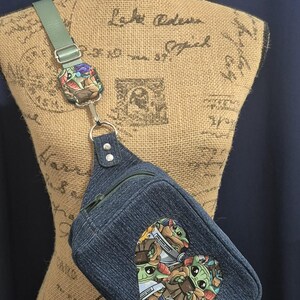 May include: Denim sling bag with a heart-shaped patch featuring a cartoon character design. The bag has a green strap and a silver-tone metal ring. The bag is displayed on a mannequin.