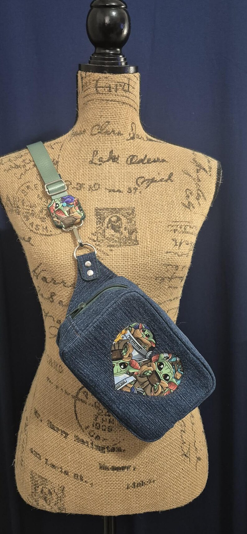 May include: Denim sling bag with a heart-shaped patch featuring a cartoon character design. The bag has a green strap and a silver-tone metal ring. The bag is displayed on a mannequin.