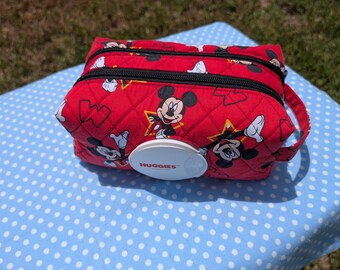 May include: A red quilted fabric pouch featuring a repeating Mickey Mouse pattern and yellow star accents. The rectangular case includes two black zippered compartments and a white plastic Huggies branded dispenser lid on the front face.