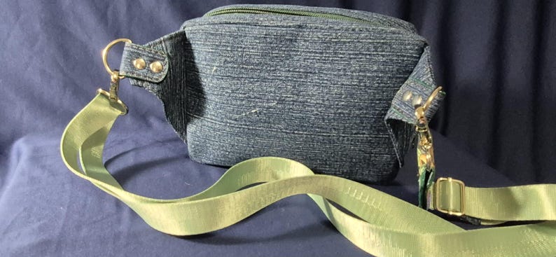 May include: A denim fanny pack with a light green strap and gold-colored hardware. The fanny pack has a zipper closure and is made of a blue denim material. The strap is adjustable and has a buckle closure. The bag is designed to be worn around the waist or across the body.