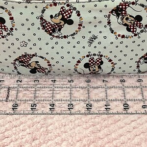 Puede incluir: Primer plano de una tela blanca con un patr&oacute;n repetido de un rat&oacute;n de dibujos animados con lunares rojos y detalles florales. La tela se mide con una regla, mostrando que tiene 17 pulgadas de largo.