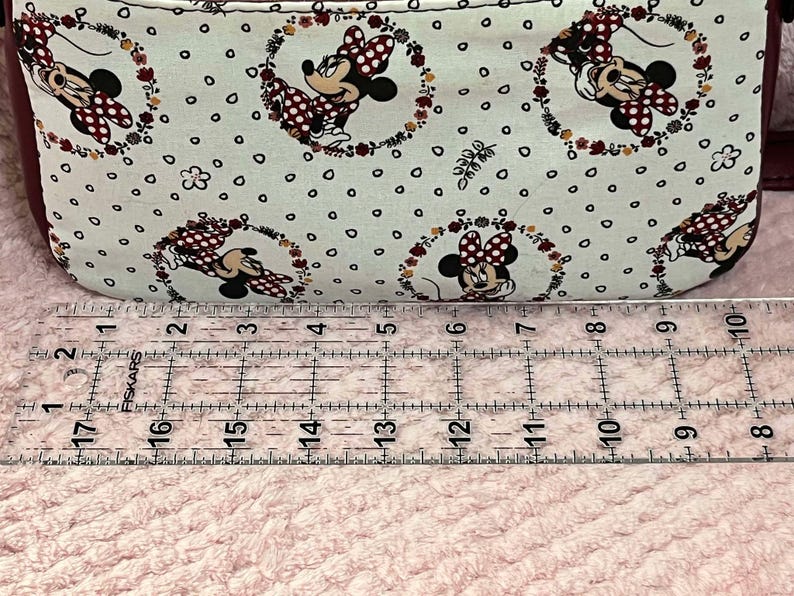 Puede incluir: Primer plano de una tela blanca con un patr&oacute;n repetido de un rat&oacute;n de dibujos animados con lunares rojos y detalles florales. La tela se mide con una regla, mostrando que tiene 17 pulgadas de largo.