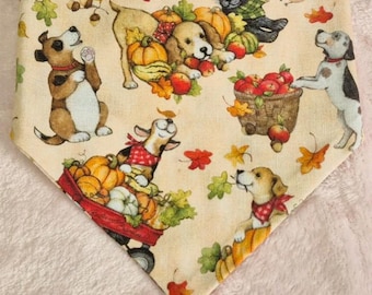 May include: Cream-colored dog bandana with a fall-themed print. The design features cartoon dogs, pumpkins, apples, and autumn leaves. The bandana is triangular in shape and made of fabric.