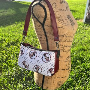 Puede incluir: Un peque&ntilde;o bolso bandolera blanco y rojo con estampado de Minnie Mouse. El bolso tiene una correa burdeos y un cierre de cremallera.