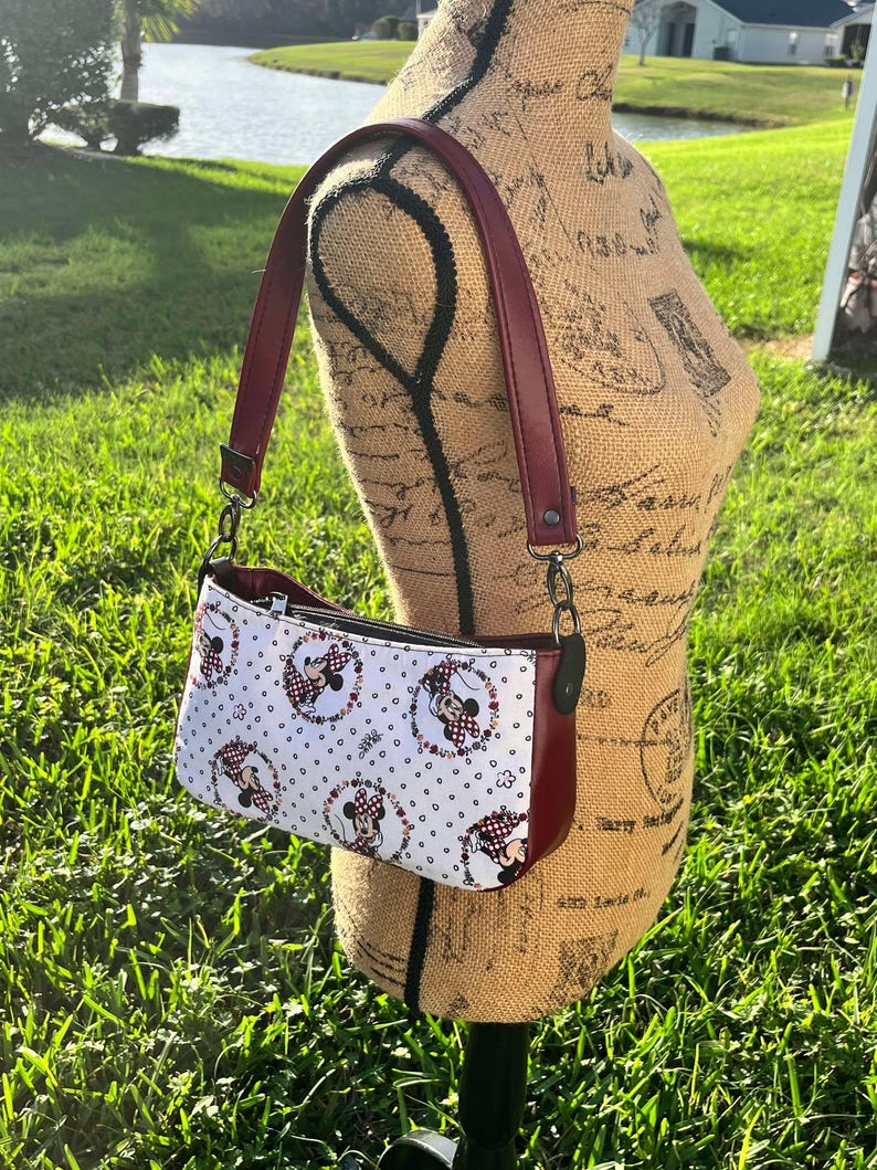Puede incluir: Un peque&ntilde;o bolso bandolera blanco y rojo con estampado de Minnie Mouse. El bolso tiene una correa burdeos y un cierre de cremallera.