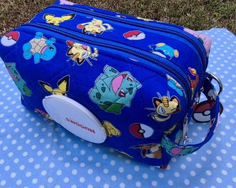 May include: A rectangular blue quilted fabric pouch featuring a repeating pattern of various colorful cartoon characters and red and white spheres. The pouch includes a white circular plastic dispenser lid with the red text Huggies printed on it.