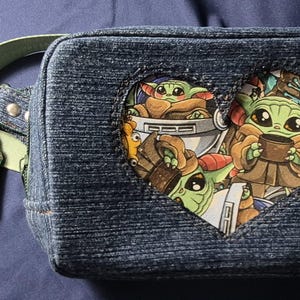 May include: Denim fanny pack with a heart-shaped cutout featuring Grogu from The Mandalorian. The pack has a light green strap and a matching charm. The main design showcases Grogu in various poses.