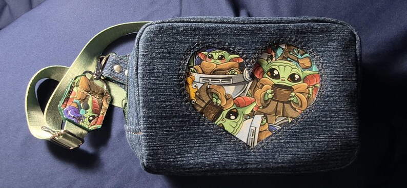 May include: Denim fanny pack with a heart-shaped cutout featuring Grogu from The Mandalorian. The pack has a light green strap and a matching charm. The main design showcases Grogu in various poses.