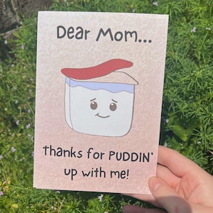 May include: A pink card with a cartoon illustration of a container of pudding with a red lid and a smiling face. The text on the card reads "Dear Mom... thanks for PUDDIN' up with me!"