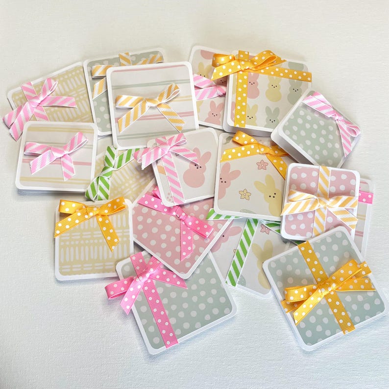 May include: A collection of square gift tags with various patterns, including stripes, polka dots, and bunny prints. Each tag is adorned with a colorful ribbon bow in shades of pink, yellow, and green. The tags are scattered on a white surface.