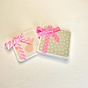 May include: Two square gift boxes with pink ribbons. One box has a light green background with white polka dots. The other box has a bunny graphic and pink and white striped ribbon.