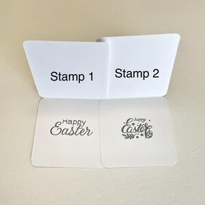 May include: Two white stamp pads labeled "Stamp 1" and "Stamp 2" with "Happy Easter" stamps. One stamp has cursive text, the other has text and floral elements. The stamps are for Easter crafts.