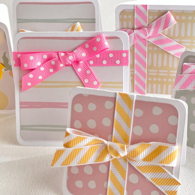 May include: Square gift boxes with rounded corners, decorated with patterned paper and ribbons. The boxes feature pink, yellow, and green stripes and polka dots. Each box is tied with a matching ribbon bow, creating a festive and decorative look.