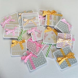 May include: A collection of square gift tags with various patterns and ribbons. Designs include polka dots, stripes, and bunny prints. Ribbons are in shades of pink, yellow, and green. The tags are arranged on a white surface.