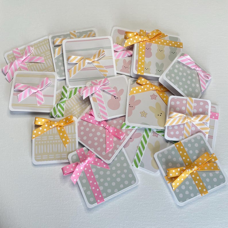 May include: A collection of square gift tags with various patterns and ribbons. Designs include polka dots, stripes, and bunny prints. Ribbons are in shades of pink, yellow, and green. The tags are arranged on a white surface.