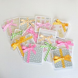 May include: A collection of square gift tags with colorful patterned designs and decorative bows. The tags feature pastel colors, polka dots, stripes, and Easter-themed patterns. The bows are in shades of pink, yellow, and green.