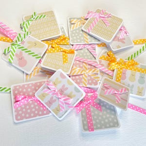 May include: A collection of square gift tags with pastel patterns and decorative ribbons. The tags feature polka dots, bunny illustrations, and geometric designs. Ribbons are pink, yellow, and green with polka dots and stripes.