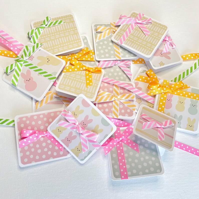 May include: A collection of square gift tags with pastel patterns and decorative ribbons. The tags feature polka dots, bunny illustrations, and geometric designs. Ribbons are pink, yellow, and green with polka dots and stripes.