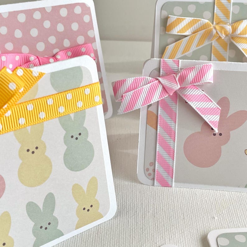 May include: Close-up of several Easter-themed gift tags. The tags feature pastel colors, polka dots, and bunny illustrations. Each tag is adorned with a decorative ribbon in various colors, including pink, yellow, and white.