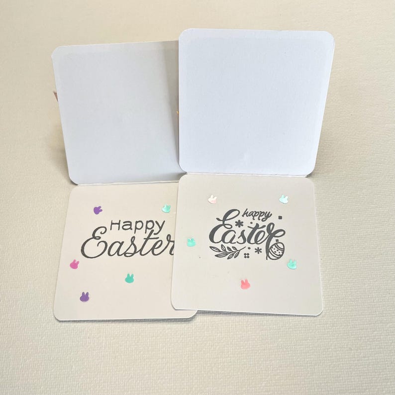 May include: Two square Easter cards with the words "Happy Easter" in black script. One card has a simple font, the other has a decorative font. Both cards have colorful bunny and confetti accents. The cards are open, revealing blank white interiors.