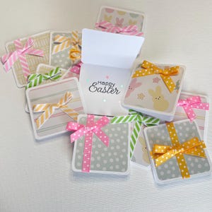 May include: A collection of square Easter cards with decorative bows. The cards feature pastel colors, polka dots, and bunny patterns. One card is open, displaying the text "Happy Easter". The bows are pink, yellow, and green.