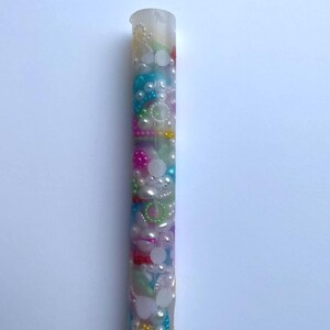 May include: A clear, cylindrical resin pen filled with colorful beads and pearls. The pen features a mix of white, pink, blue, and green beads, creating a decorative and unique writing instrument. The pen is approximately 6 inches long.