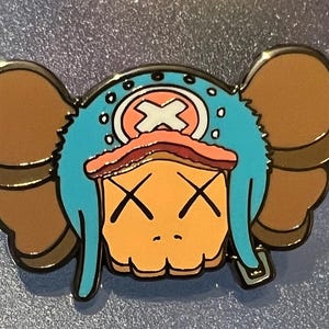 May include: A blue and brown enamel pin featuring a cartoon character with a blue hat, brown ears, and a white X on the hat. The character has a light orange face with two black X's for eyes.